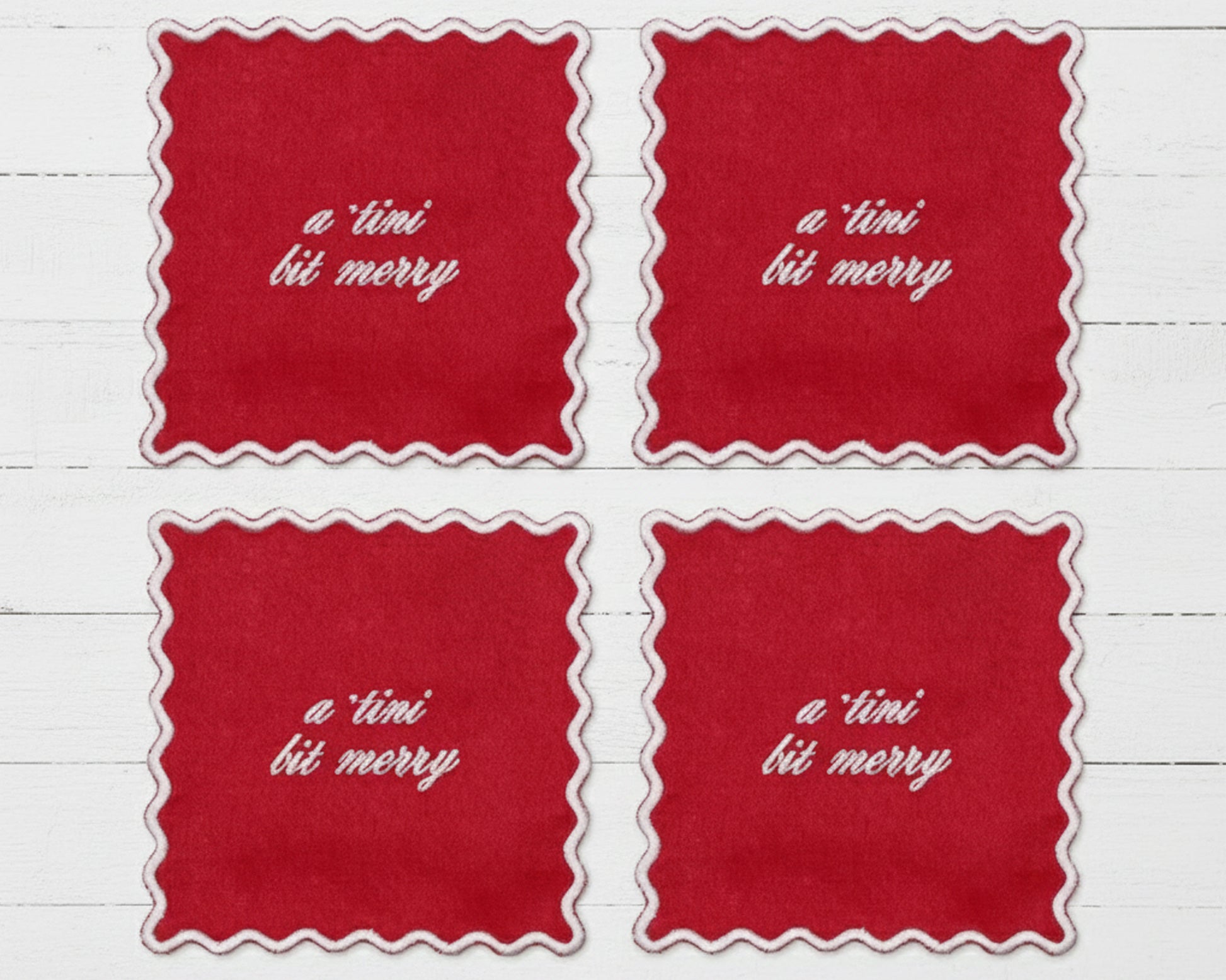 A set of four ruby red linen cocktail napkins with white scalloped edges featuring "a tini bit merry" in white vinyl script.