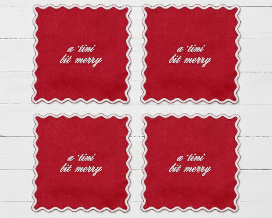 A set of four ruby red linen cocktail napkins with white scalloped edges featuring "a tini bit merry" in white vinyl script.