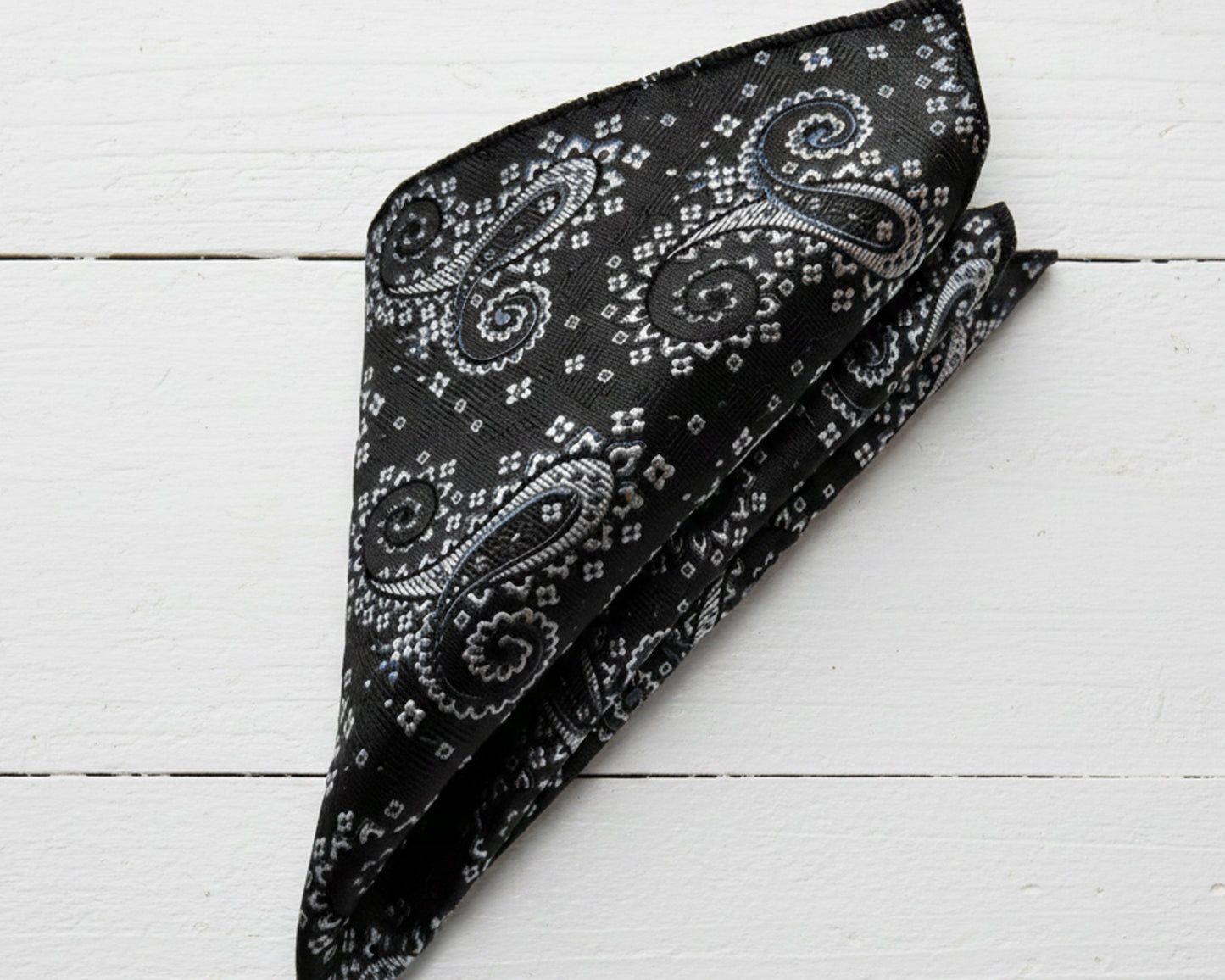 Close up of a midnight onyx paisley cotton handkerchief styled in a classic puff fold on a luxury champagne satin surface.