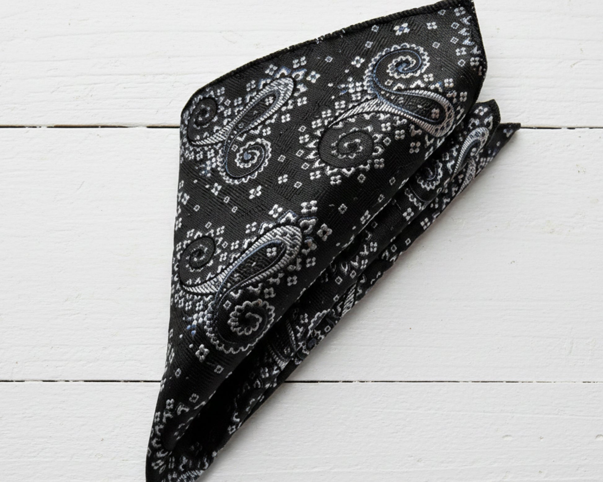 Close up of a midnight onyx paisley cotton handkerchief styled in a classic puff fold on a luxury champagne satin surface.