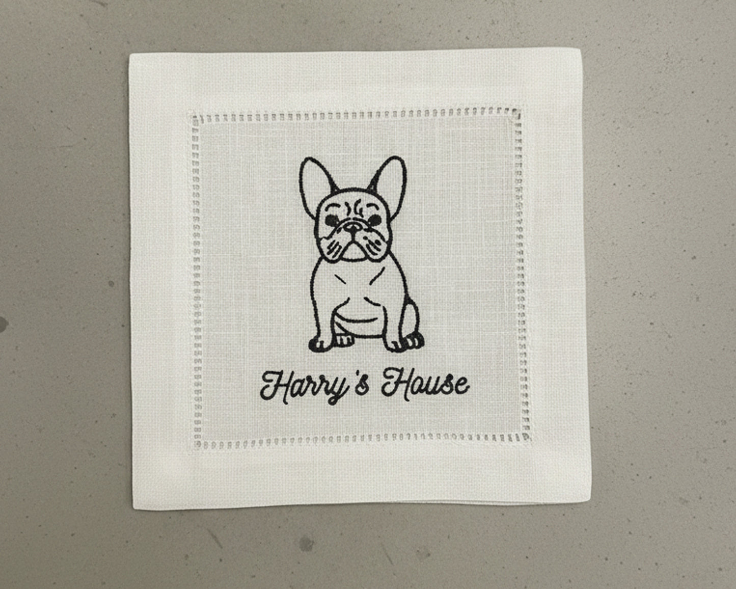 A single white 100% cotton hemstitch napkin featuring a detailed black machine-embroidered French Bulldog illustration and "Harry's House" artisanal script.