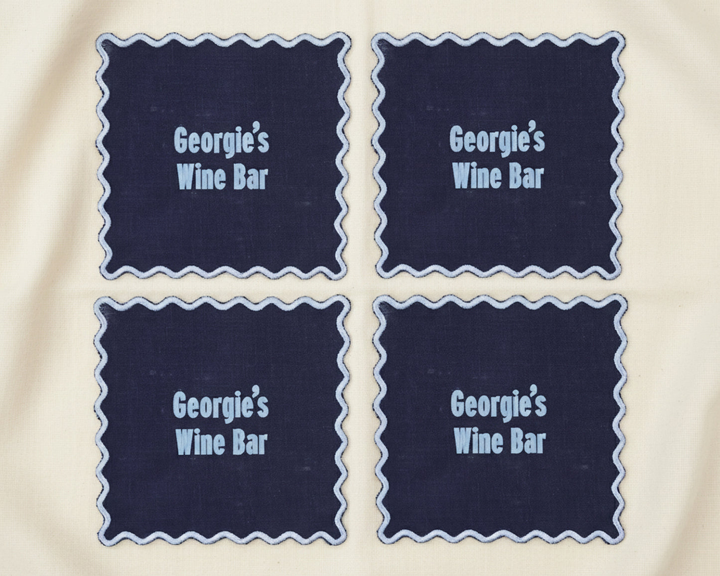A set of four navy blue linen cocktail napkins with white scalloped edges featuring "Georgie’s Wine Bar" in light blue vinyl.
