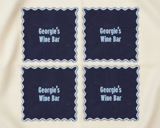 A set of four navy blue linen cocktail napkins with white scalloped edges featuring "Georgie’s Wine Bar" in light blue vinyl.