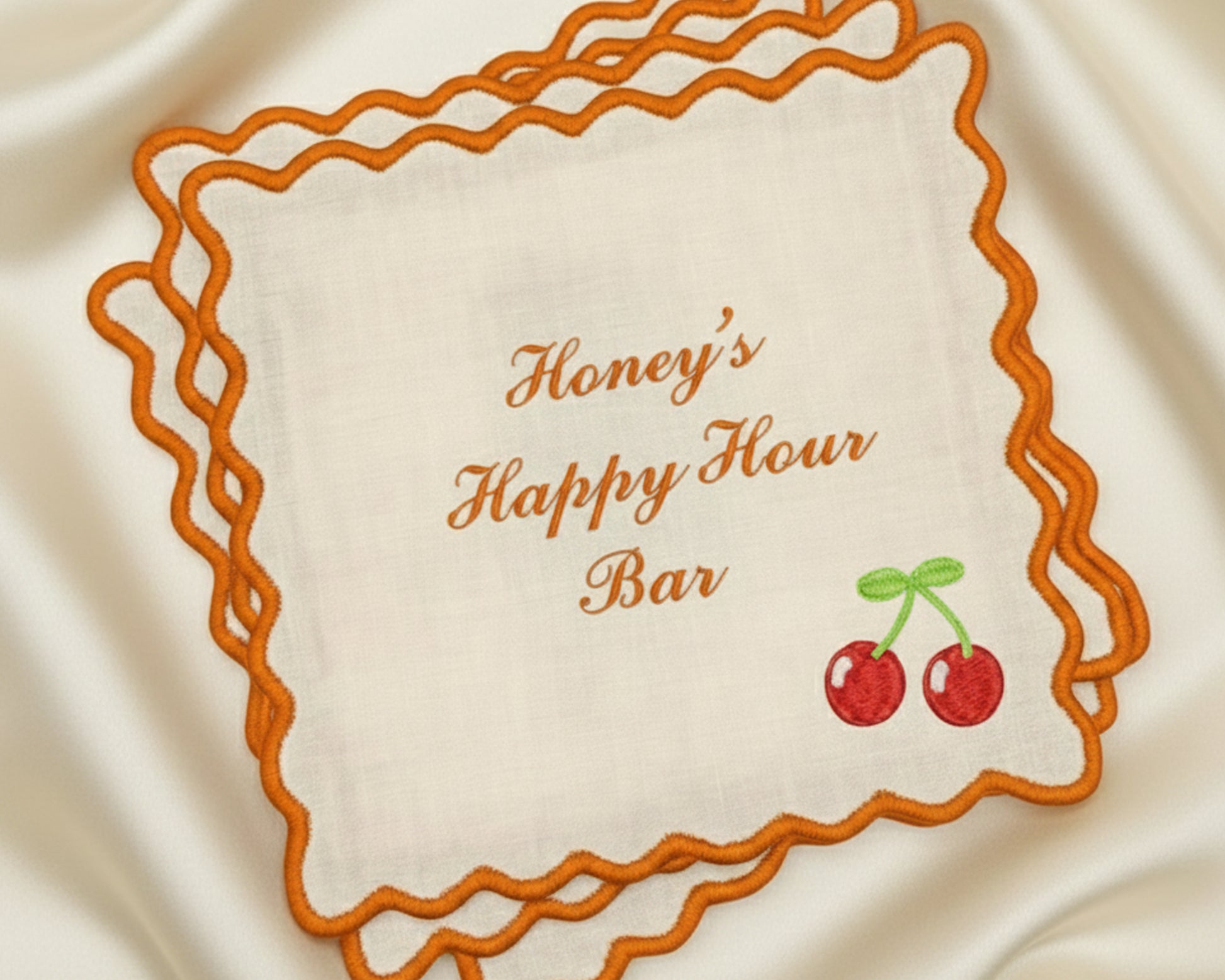 A beige cotton bar napkin with a vibrant orange scalloped border, personalized with "Honey's Happy Hour Bar" and a colorful embroidered cherry icon.