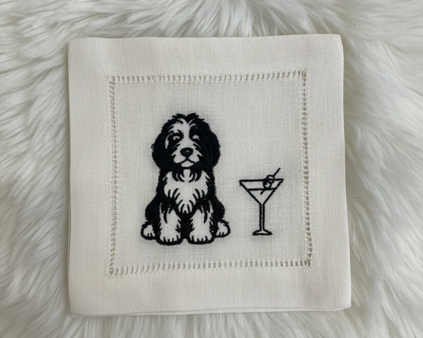 A white cotton hemstitch napkin featuring a high-density black machine-embroidered illustration of a Doodle dog sitting next to a garnished martini glass.