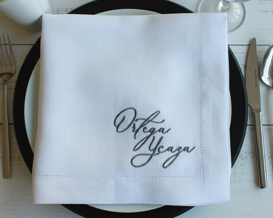 White napkin with 'Ortega Ycaza' embroidery on a table setting with cutlery.