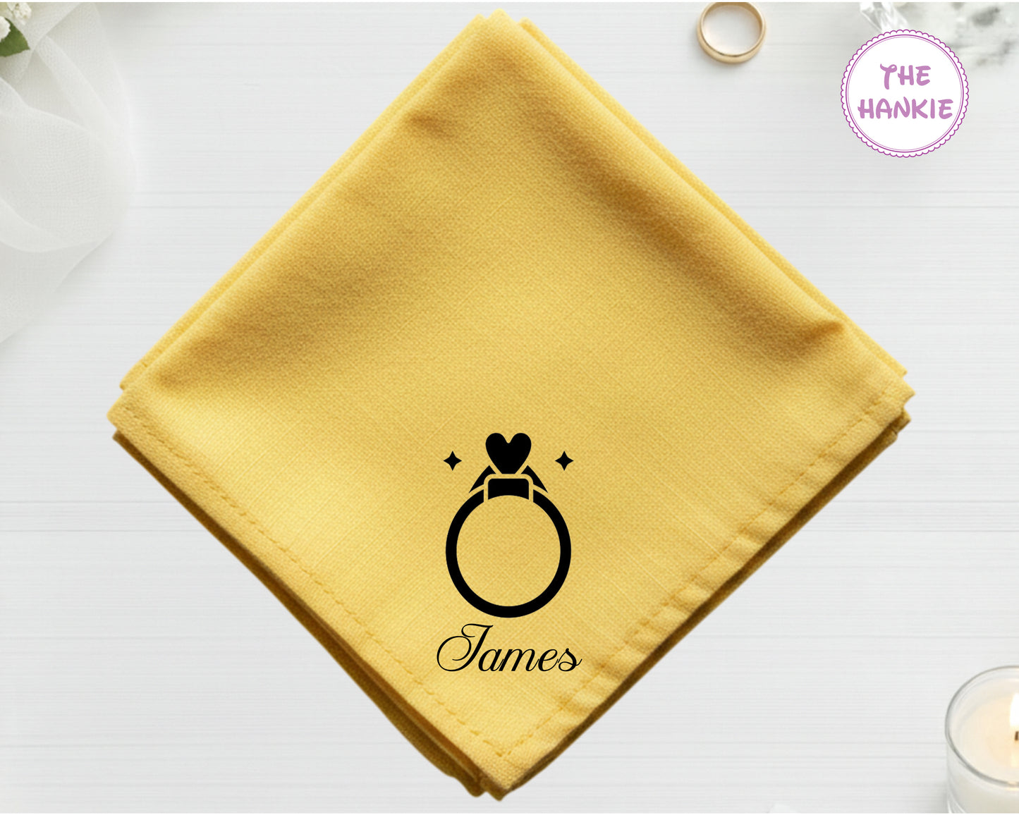 A golden harvest yellow cotton handkerchief personalized with the name "James" below an embroidered black diamond ring with a heart-shaped stone.