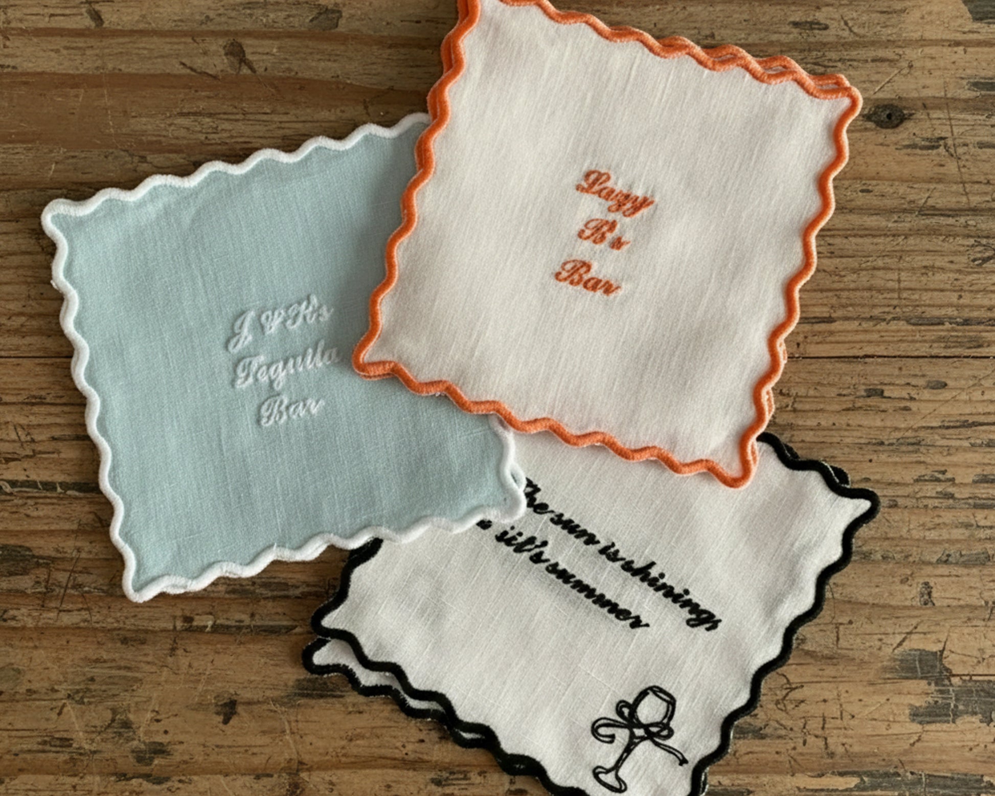 A set of white linen coasters with black and orange scalloped edges, featuring custom embroidery for "Janie's Cocktail Bar" and "Lazy B's Bar" with cocktail icons.