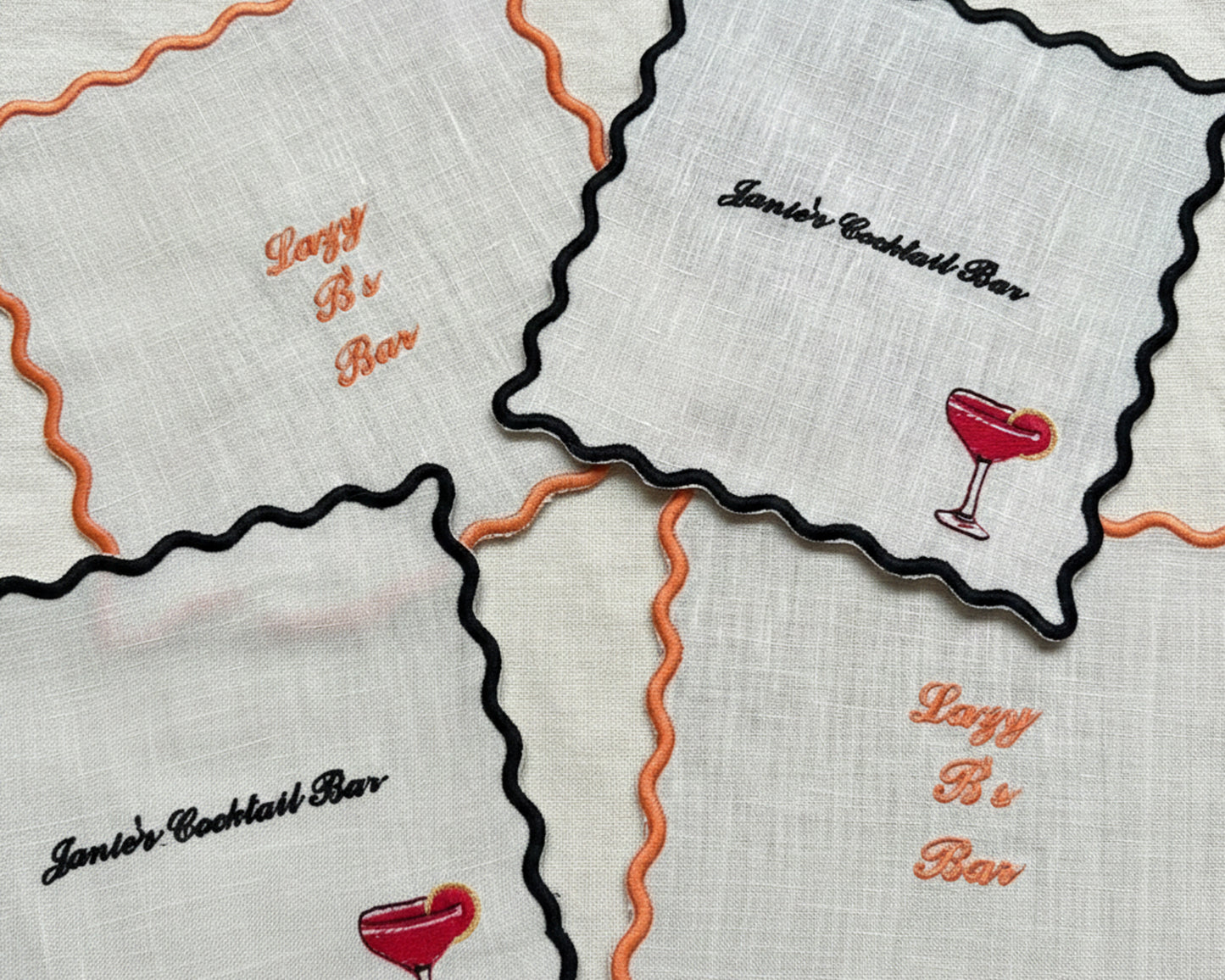 A collection of white cotton coasters with black and orange scalloped edges, featuring custom embroidery for "Janie's Cocktail Bar" and "Lazy B's Bar" with drink icons.