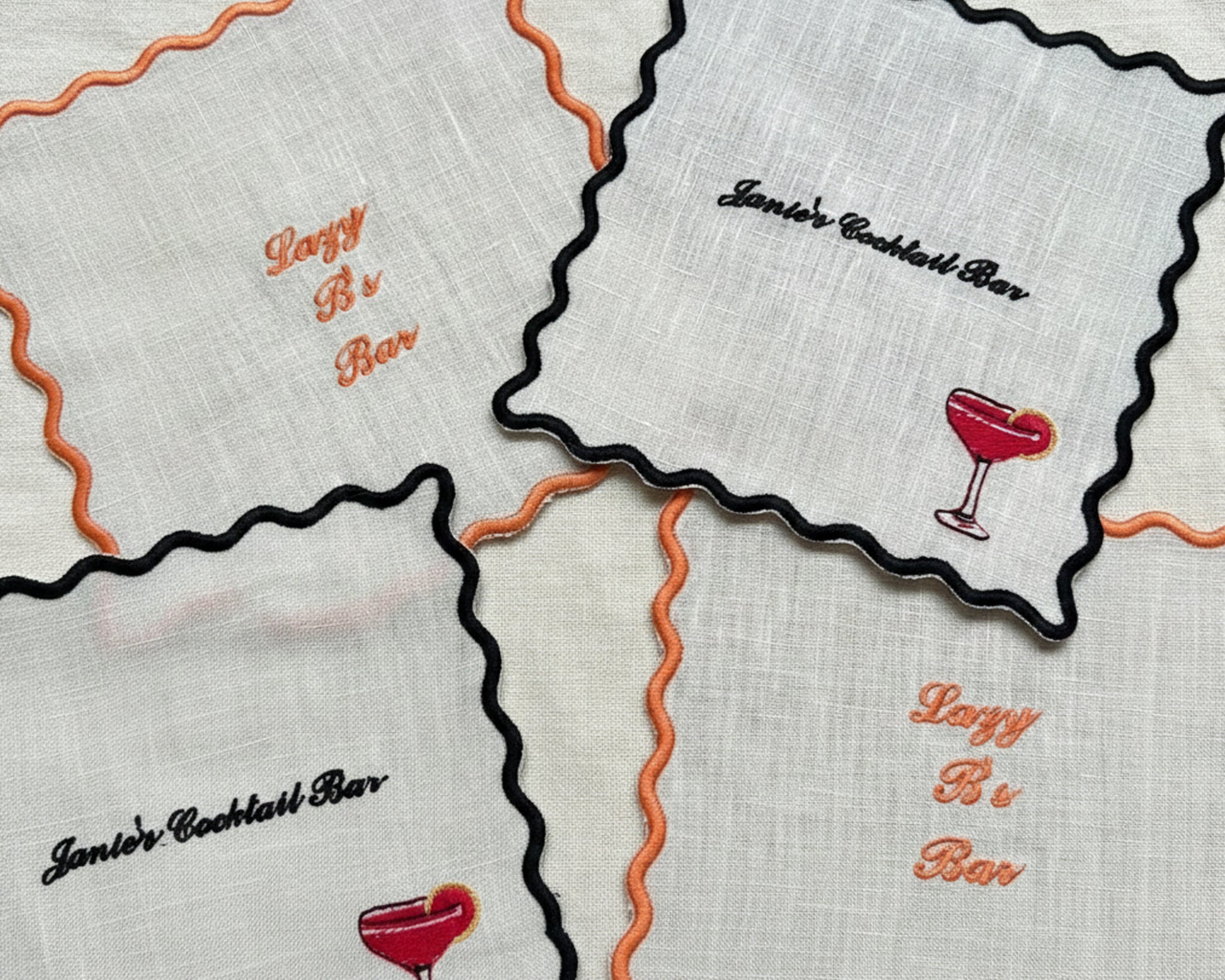 A collection of white cotton coasters with black and orange scalloped edges, featuring custom embroidery for "Janie's Cocktail Bar" and "Lazy B's Bar" with drink icons.