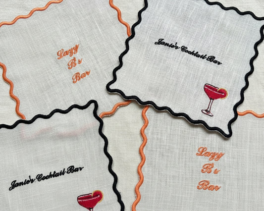 A collection of white cotton coasters with black and orange scalloped edges, featuring custom embroidery for "Janie's Cocktail Bar" and "Lazy B's Bar" with drink icons.