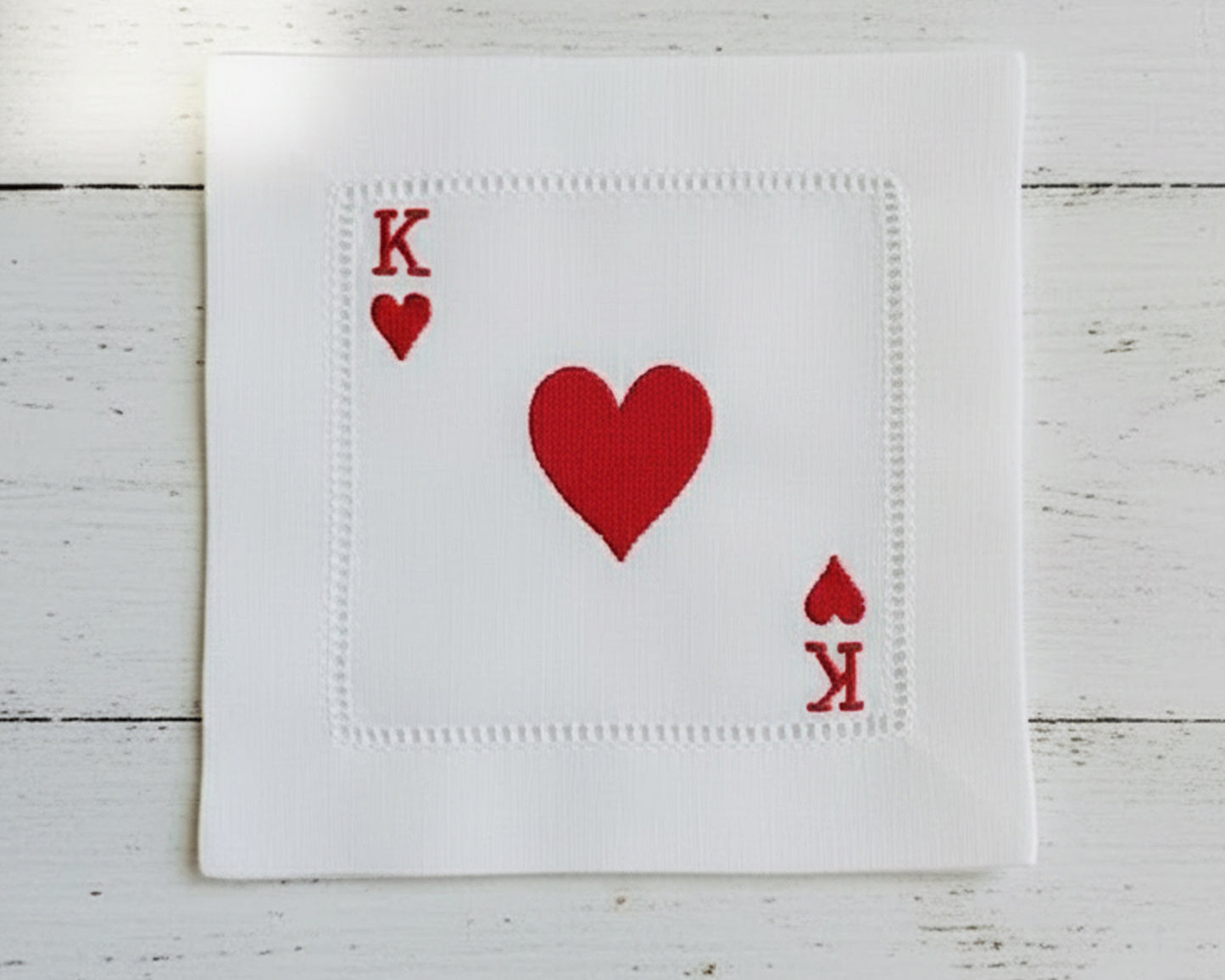 A set of three white cotton beverage napkins with hemstitch borders, featuring the Jack, Queen, and King of Hearts embroidered in red thread.