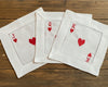 A set of three white cotton beverage napkins with hemstitch borders, featuring the Jack, Queen, and King of Hearts embroidered in red thread.