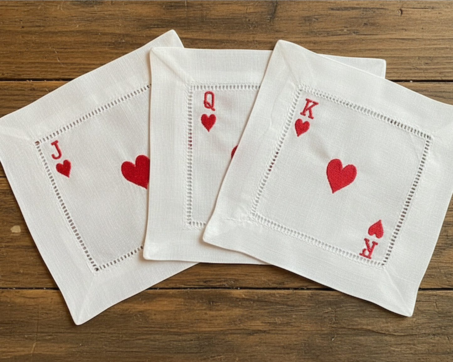 A set of three white cotton beverage napkins with hemstitch borders, featuring the Jack, Queen, and King of Hearts embroidered in red thread.