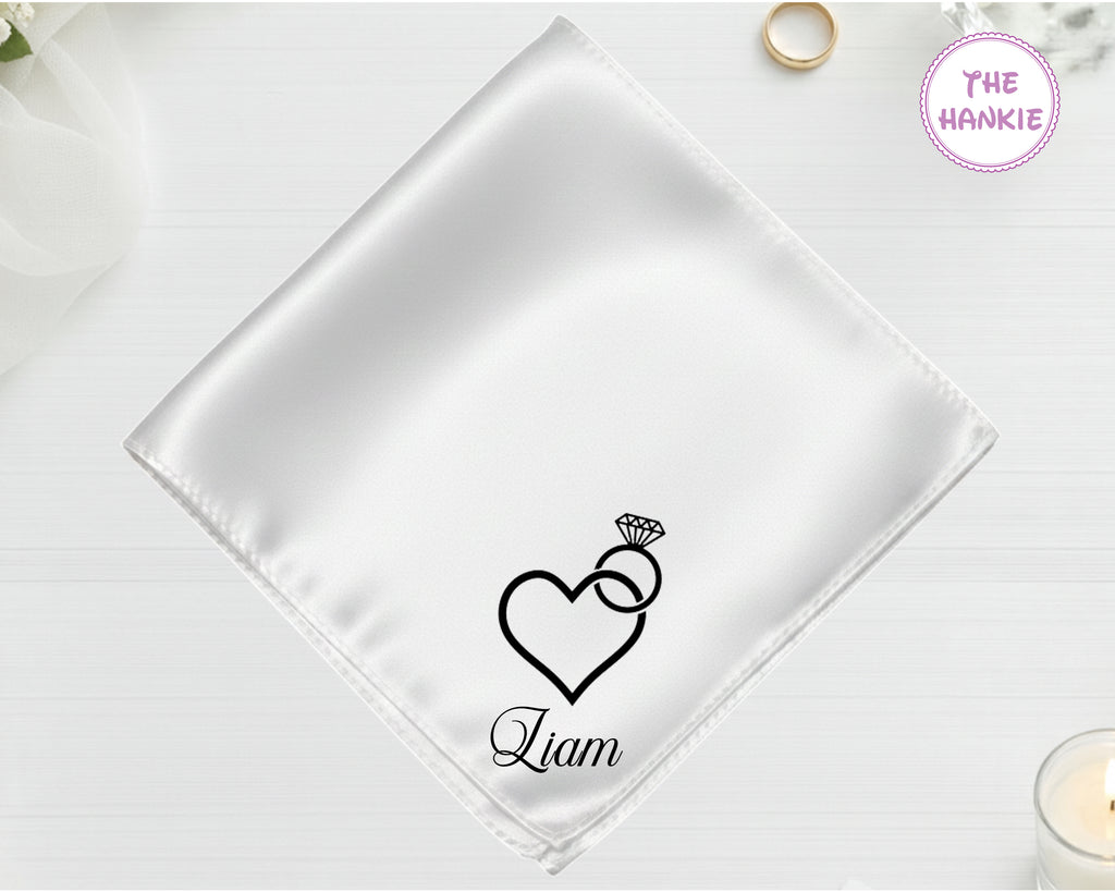 A shimmering pearl white luxury satin handkerchief personalized with the name "Liam" below an embroidered diamond ring with a heart-shaped stone.