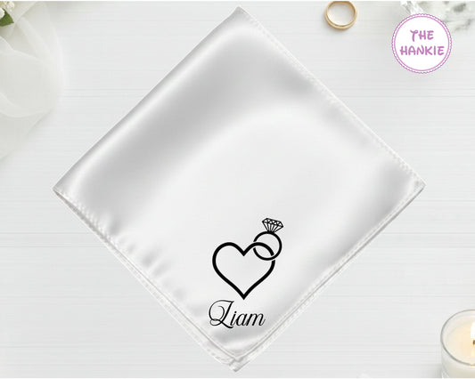 A shimmering pearl white luxury satin handkerchief personalized with the name "Liam" below an embroidered diamond ring with a heart-shaped stone.
