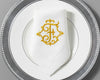 Luxury white cotton napkin with gold embroidered T monogram, folded on elegant silver-rim dinner plate