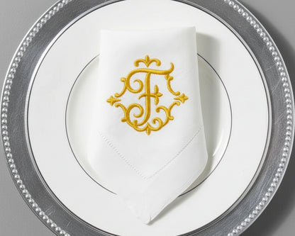 Luxury white cotton napkin with gold embroidered T monogram, folded on elegant silver-rim dinner plate