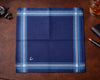 Folded blue plaid fabric on a brown leather surface with a glass of whiskey and accessories.