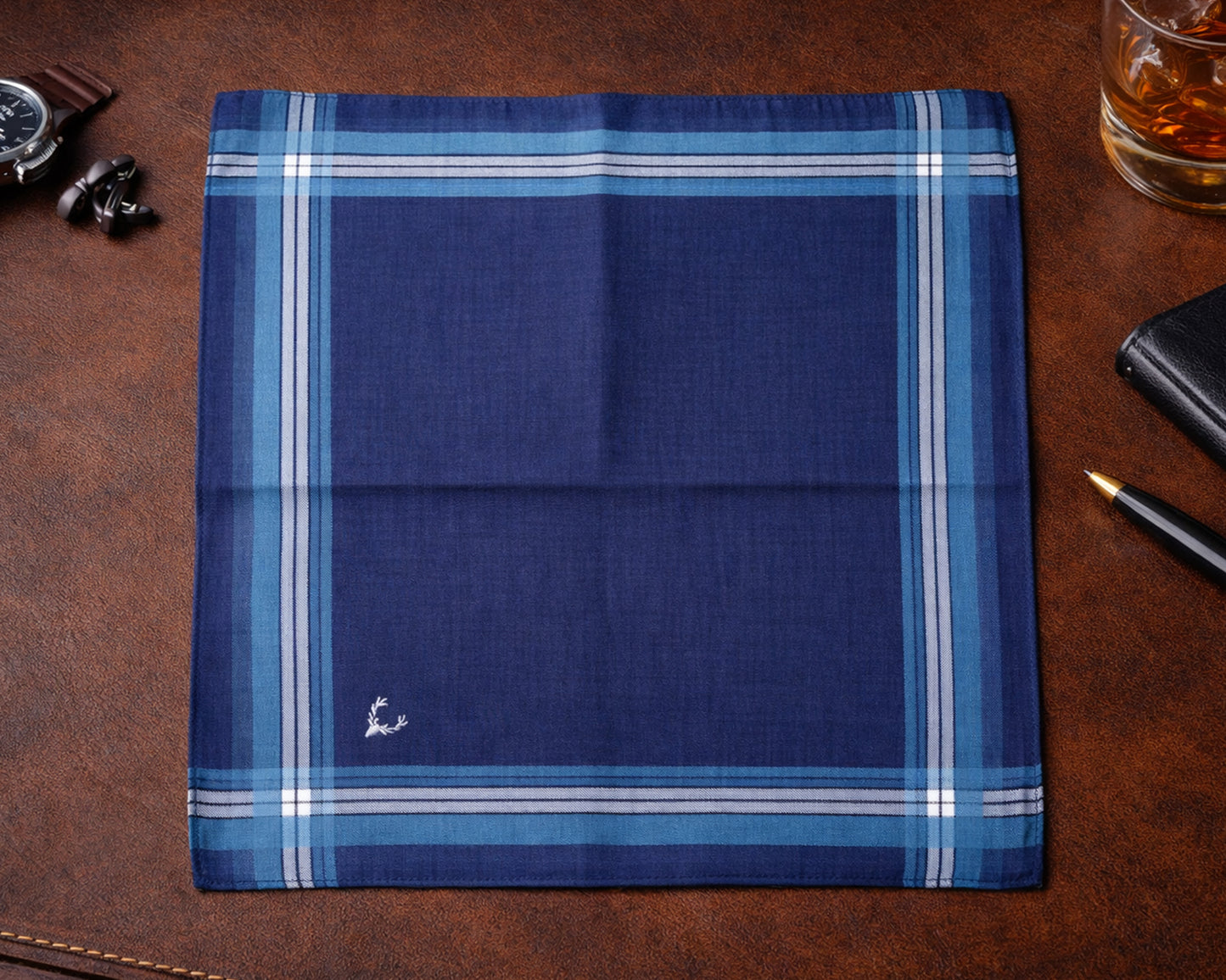 Folded blue plaid fabric on a brown leather surface with a glass of whiskey and accessories.