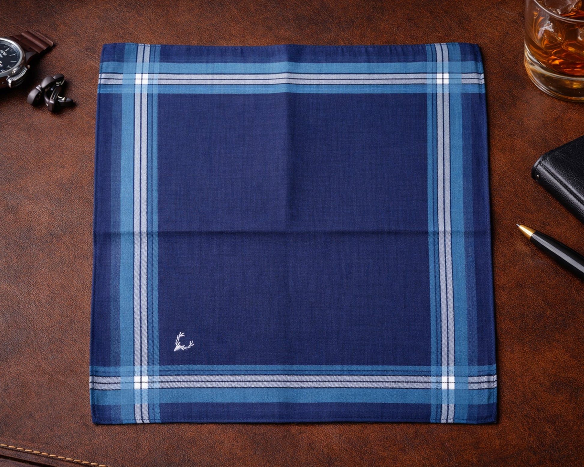 Folded blue plaid fabric on a brown leather surface with a glass of whiskey and accessories.