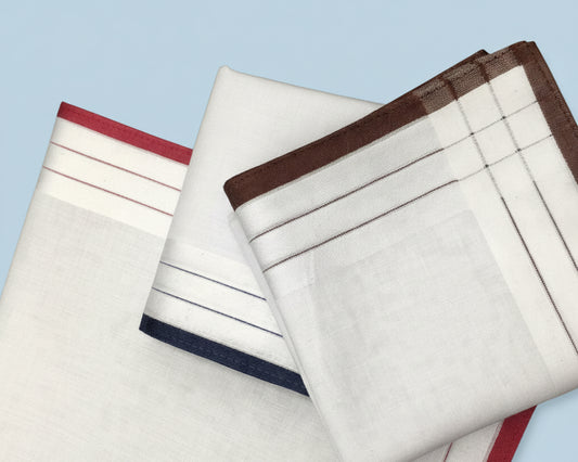 Flat lay of three white cotton handkerchiefs featuring red, navy, and brown woven striped borders on a light blue background.