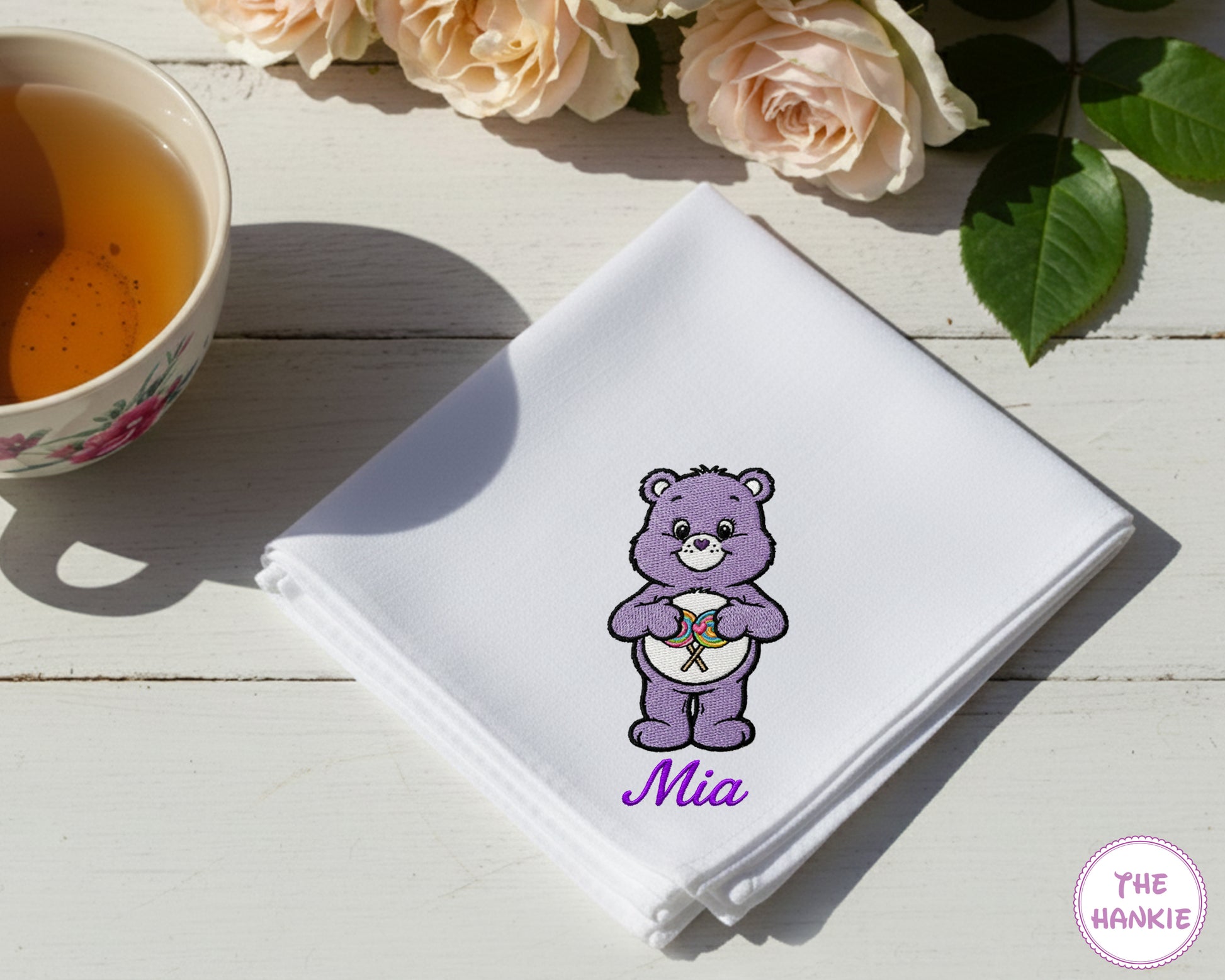 A premium white cotton handkerchief customized with the name "Mia" in purple script and an embroidered purple Share Bear character holding lollipops.