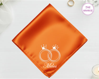 A radiant sunset orange high-lustre satin pocket square featuring the name "Miles" and white embroidery of two interlocking wedding bands topped with crowns.