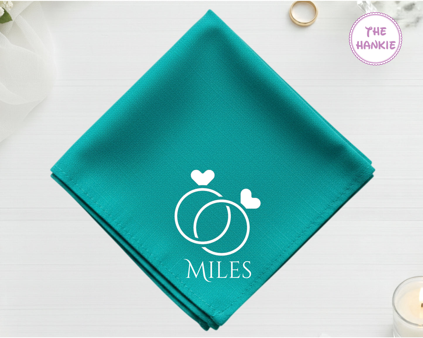 A deep teal cotton handkerchief customized with the name "Miles" and white embroidery featuring two interlocking wedding bands topped with hearts.