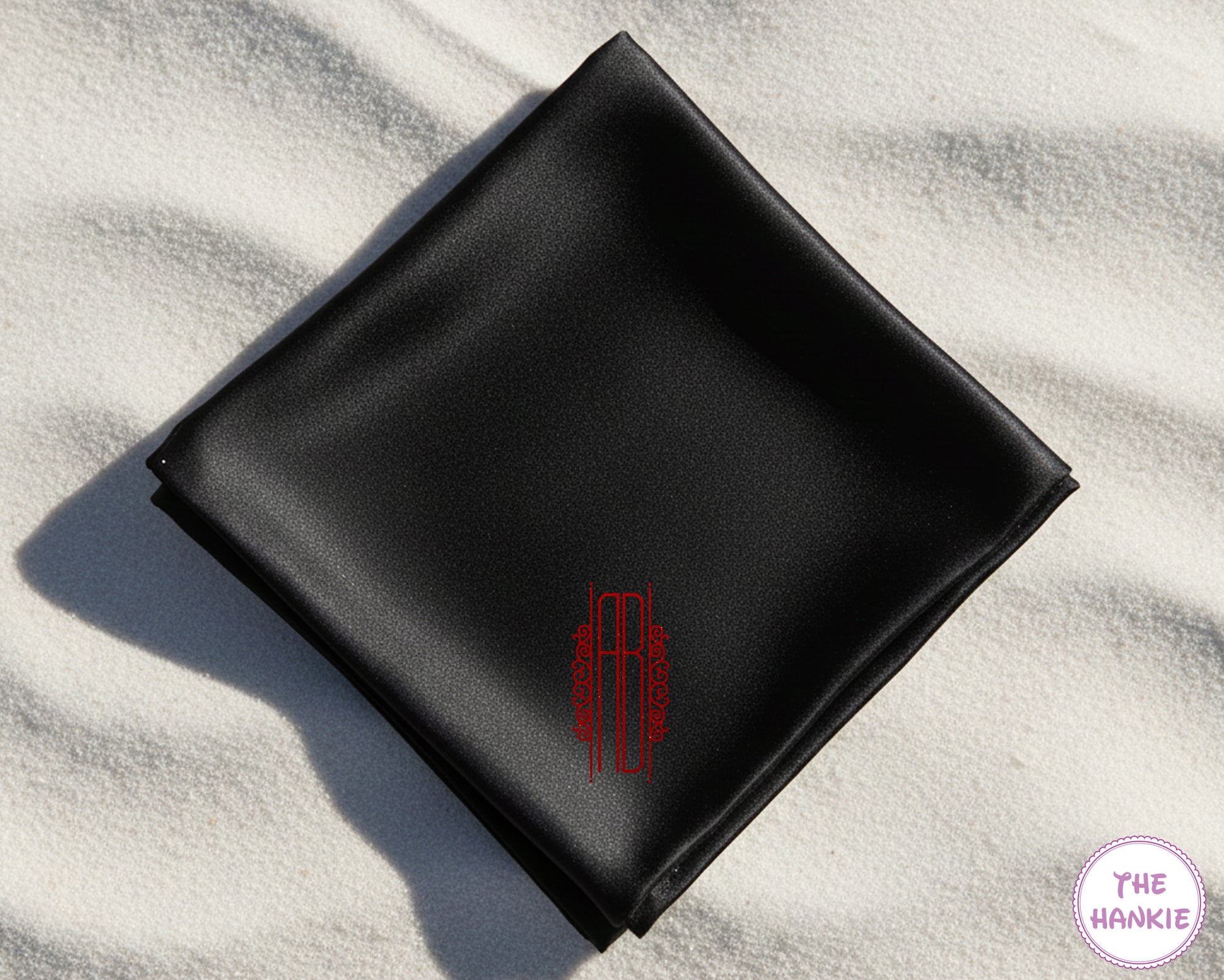 Premium black satin pocket square with custom red vinyl initials "AB" in a modern vertical script frame on a beach background.