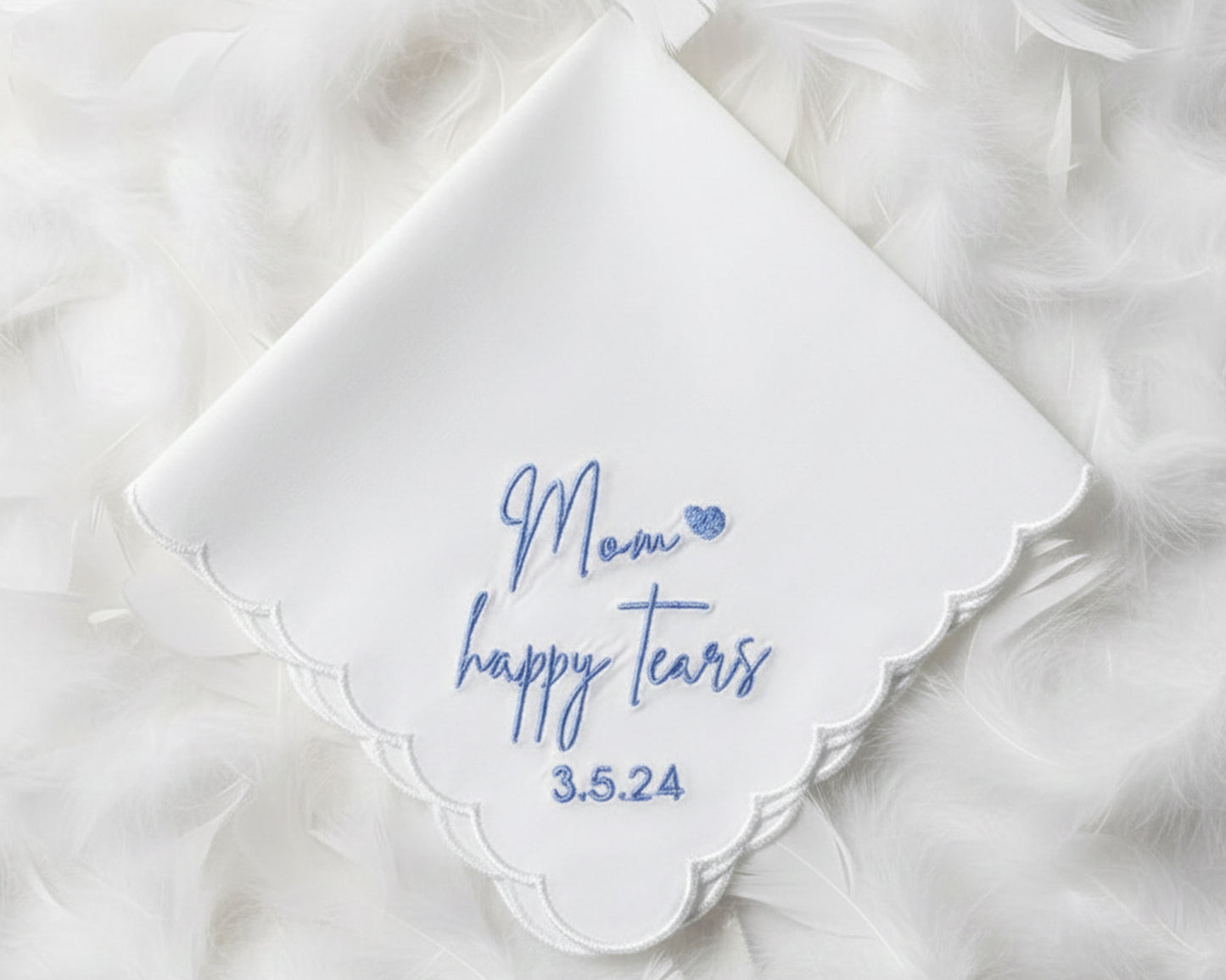 A white scalloped cotton handkerchief featuring blue "Mom happy tears" script embroidery and a wedding date, displayed on soft white feathers.