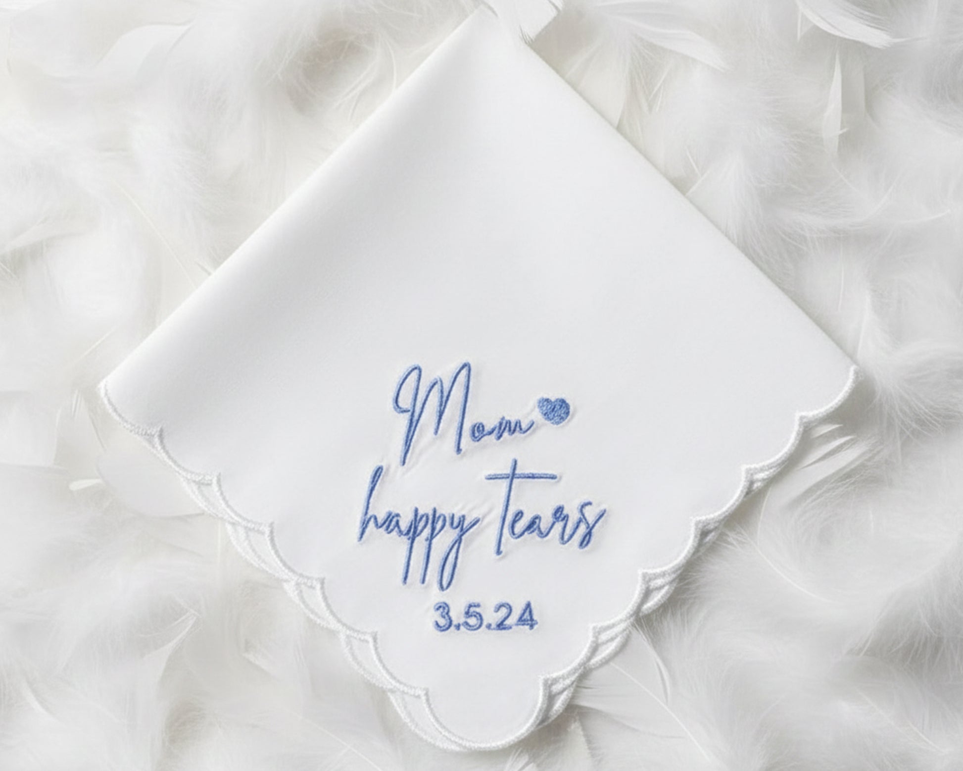 A white scalloped cotton handkerchief featuring blue "Mom happy tears" script embroidery and a wedding date, displayed on soft white feathers.