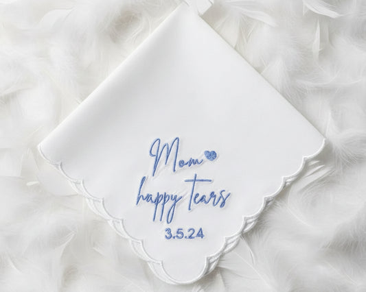 A white scalloped cotton handkerchief featuring blue "Mom happy tears" script embroidery and a wedding date, displayed on soft white feathers.