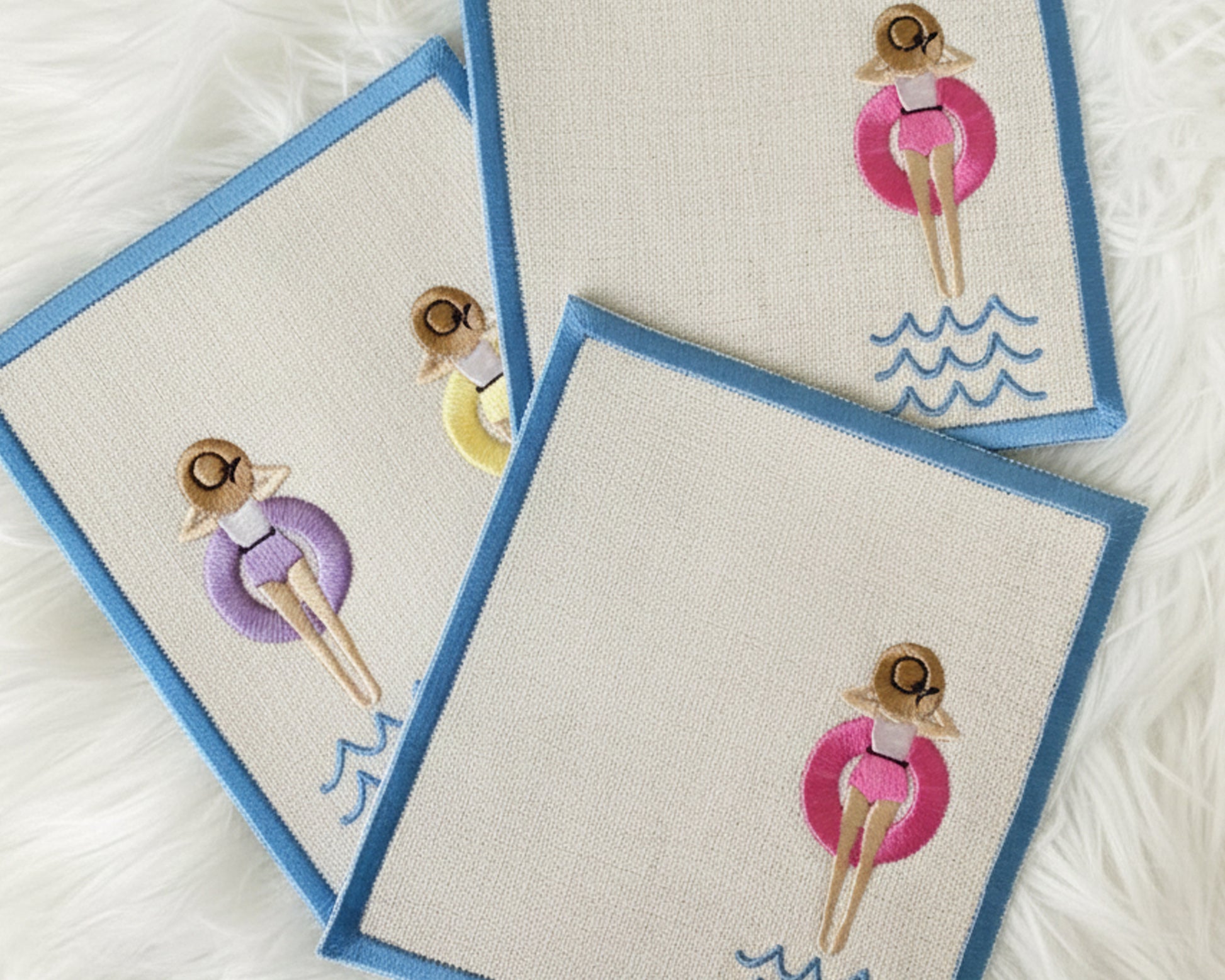 A collection of three white linen cocktail napkins with blue borders, each featuring a girl in a different colored floaty: pink, yellow, and purple.