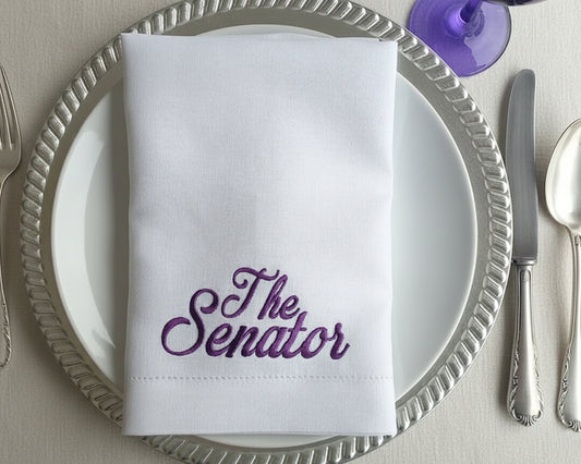 White napkin with 'The Senator' embroidered on it, placed on a silver charger plate with cutlery.