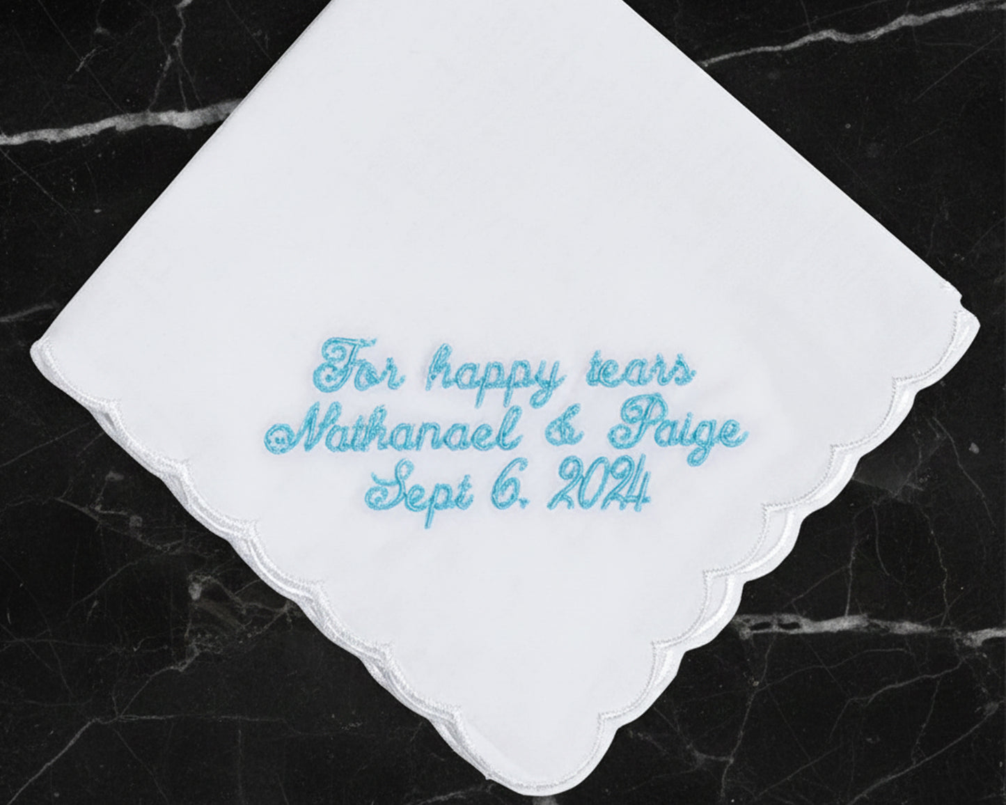 A white scalloped handkerchief featuring light blue embroidery for "For happy tears," the names Nathanael and Paige, and their wedding date.