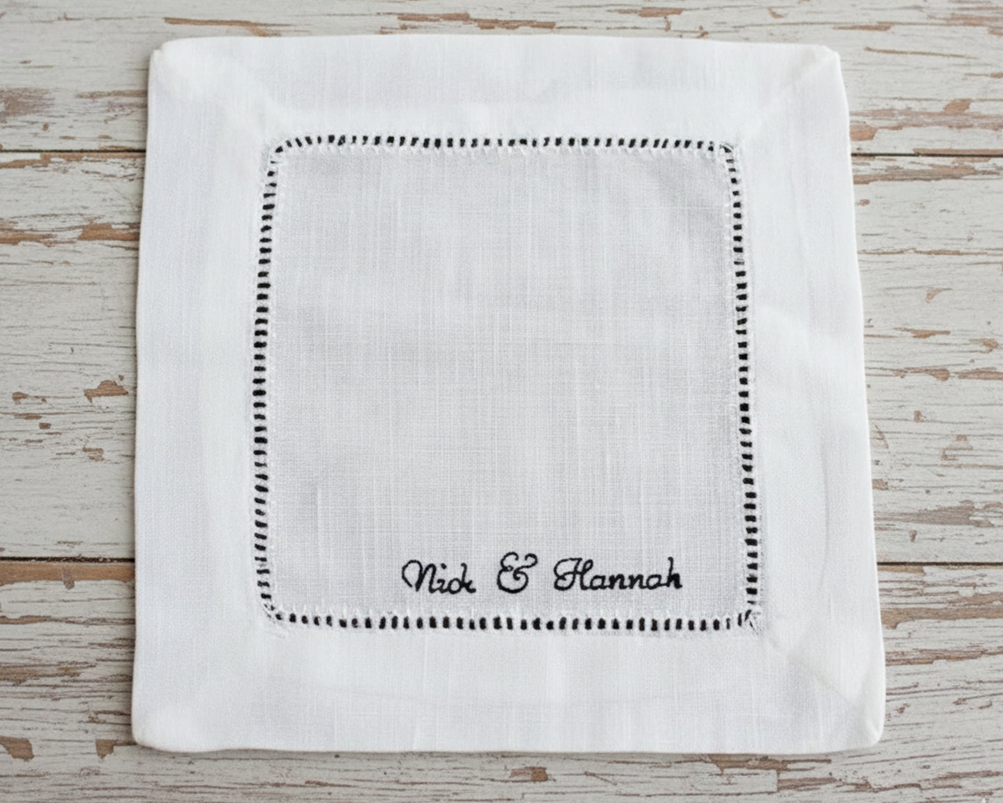 A close-up of a white linen cocktail napkin featuring a black-highlighted hemstitch border and custom "Nick & Hannah" script embroidery.