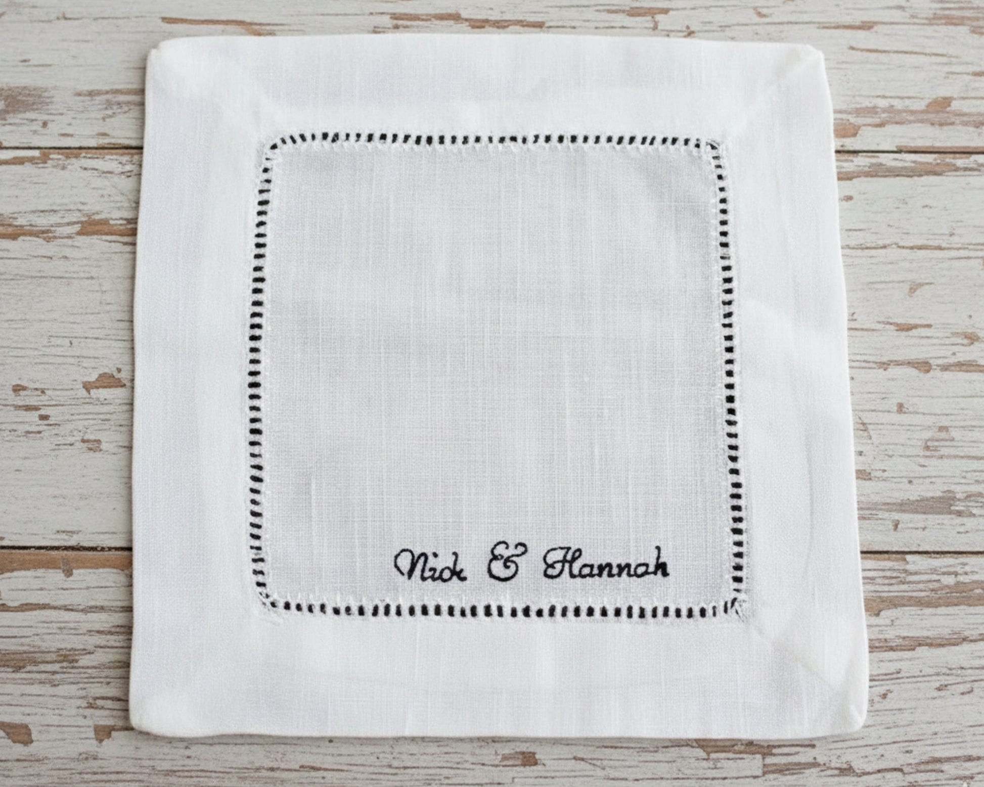 A close-up of a white linen cocktail napkin featuring a black-highlighted hemstitch border and custom "Nick & Hannah" script embroidery.
