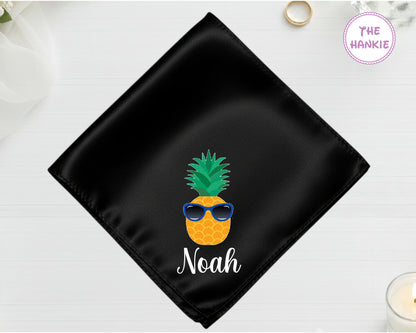 A midnight jet black high-lustre satin pocket square customized with "Noah" and a vibrant pineapple icon wearing blue-lensed sunglasses.