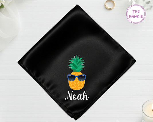 A midnight jet black high-lustre satin pocket square customized with "Noah" and a vibrant pineapple icon wearing blue-lensed sunglasses.