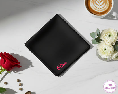 A premium black cotton handkerchief customized with the name "Oliver" in a bright pink script embroidery, shown alongside a red rose and a cup of coffee.