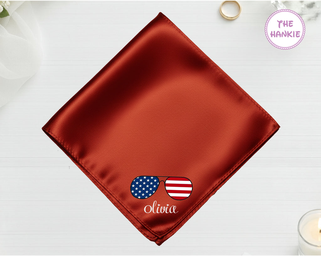 A burnished copper luxury satin handkerchief personalized with the name "Olivia" below an embroidered icon of sunglasses featuring the American flag.