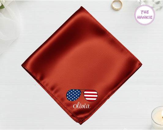 A burnished copper luxury satin handkerchief personalized with the name "Olivia" below an embroidered icon of sunglasses featuring the American flag.
