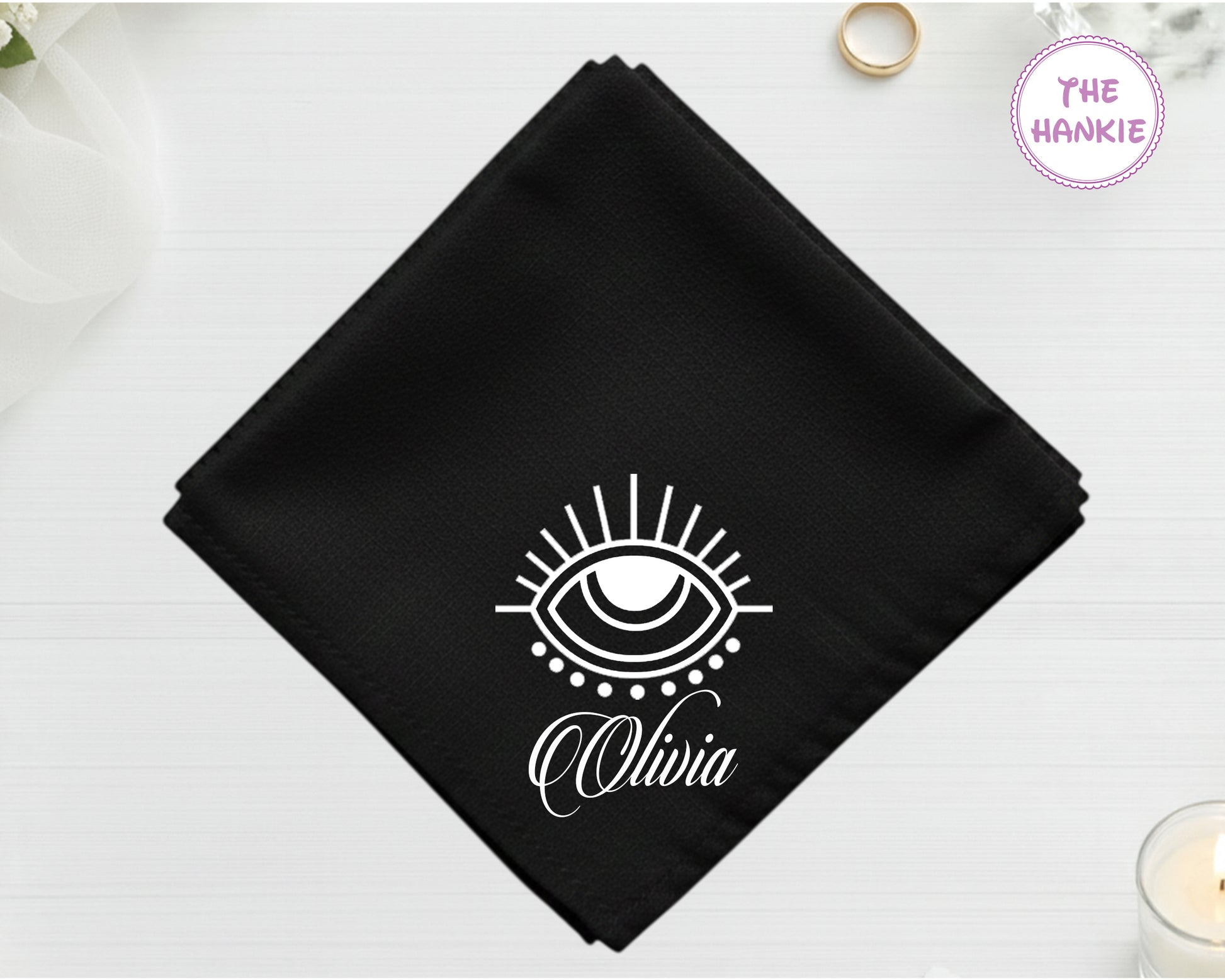 A midnight onyx black cotton pocket square featuring the name "Olivia" in white script below a minimalist white protective eye symbol.