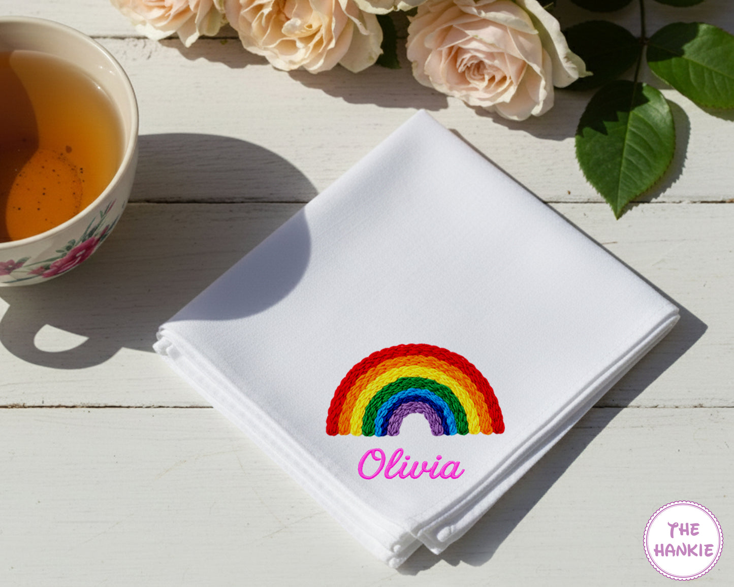 A crisp white cotton handkerchief personalized with the name "Olivia" in pink script below a vibrant, multi-color embroidered rainbow.