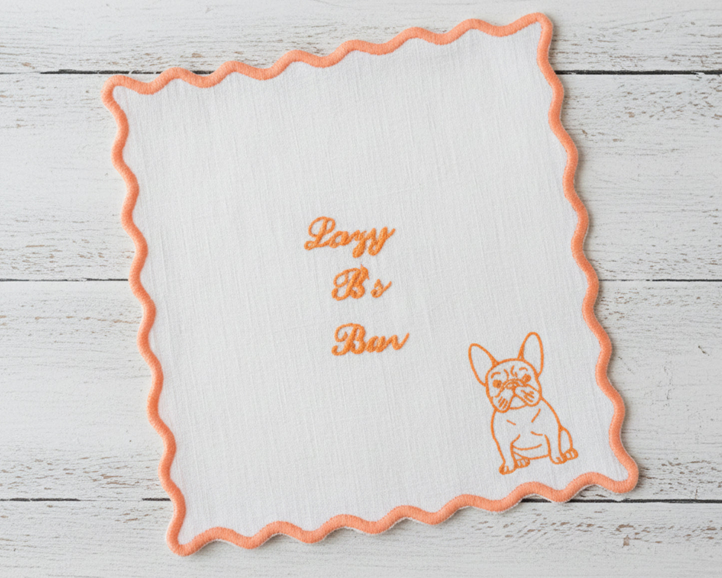 A white linen cocktail napkin with an orange scalloped edge embroidered with "Lazy B's Bar" and a Frenchie illustration.