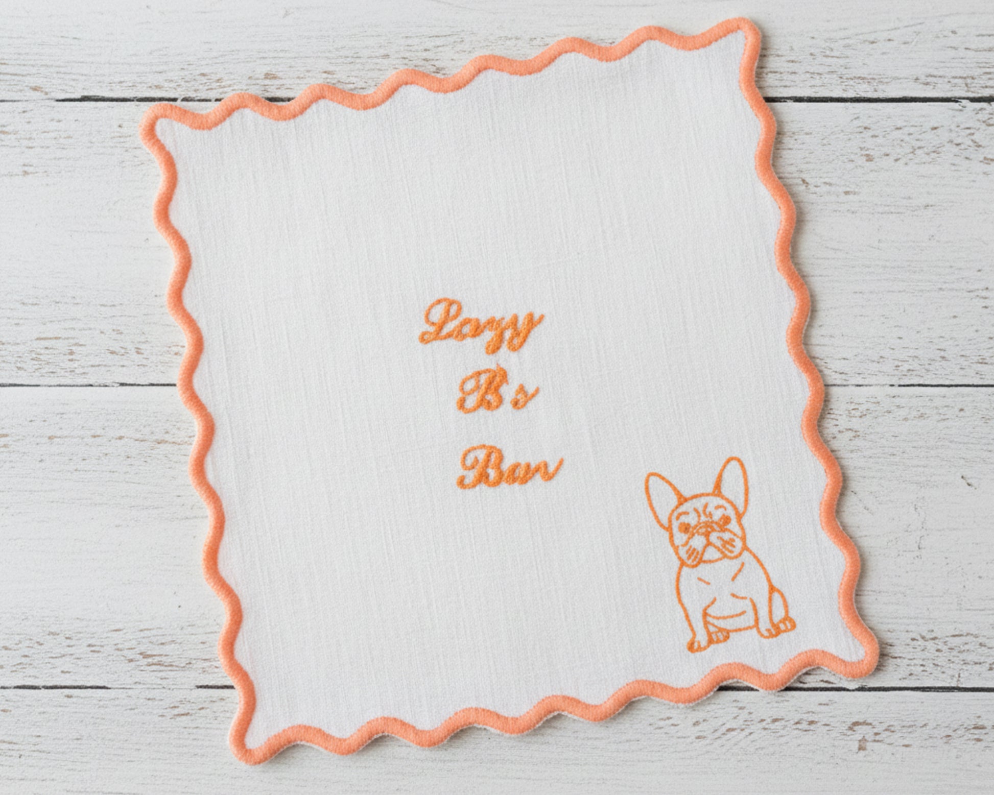 A white linen cocktail napkin with an orange scalloped edge embroidered with "Lazy B's Bar" and a Frenchie illustration.