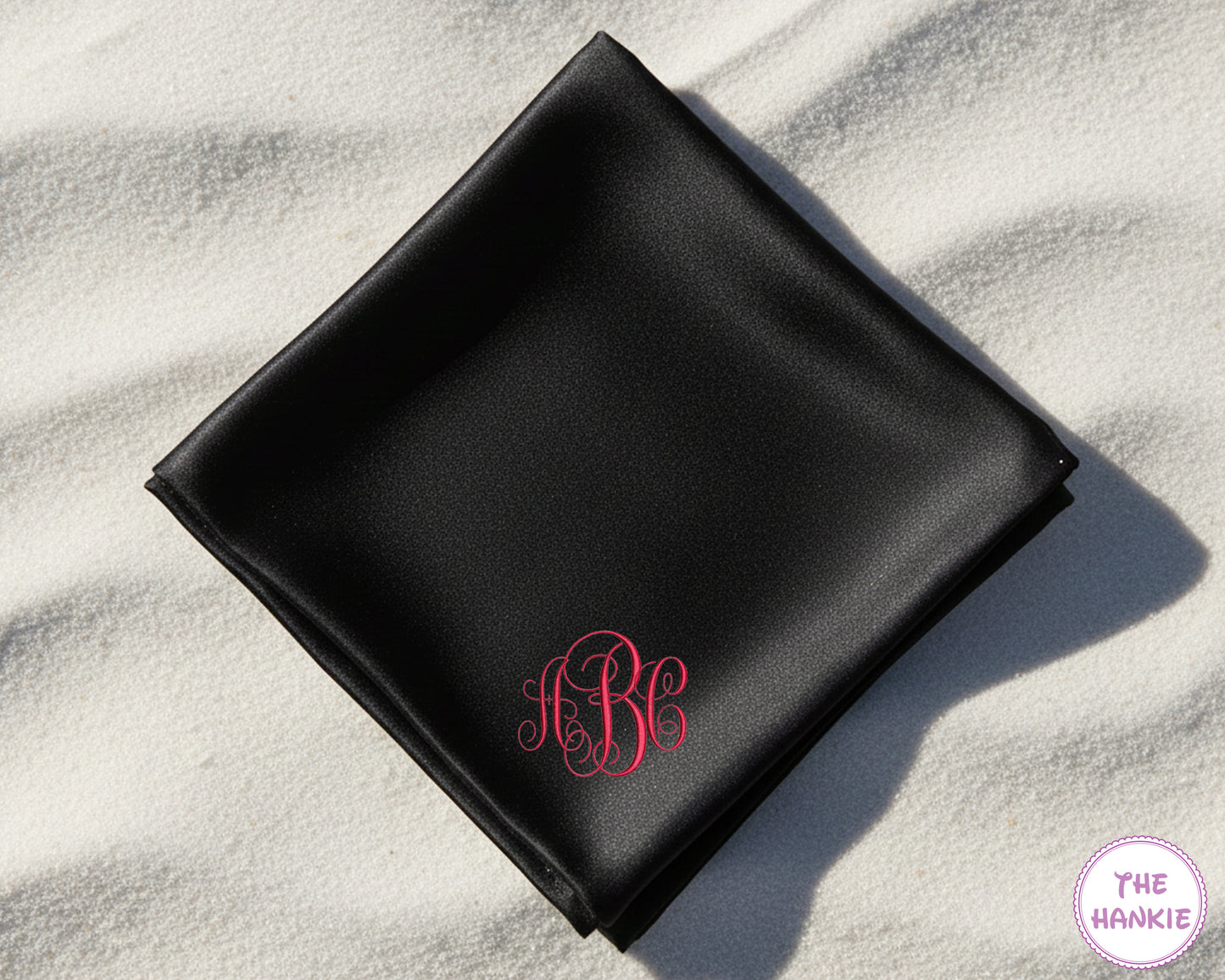 A premium black satin pocket square personalized with an ornate interlocking script monogram in a vibrant pink embroidery.