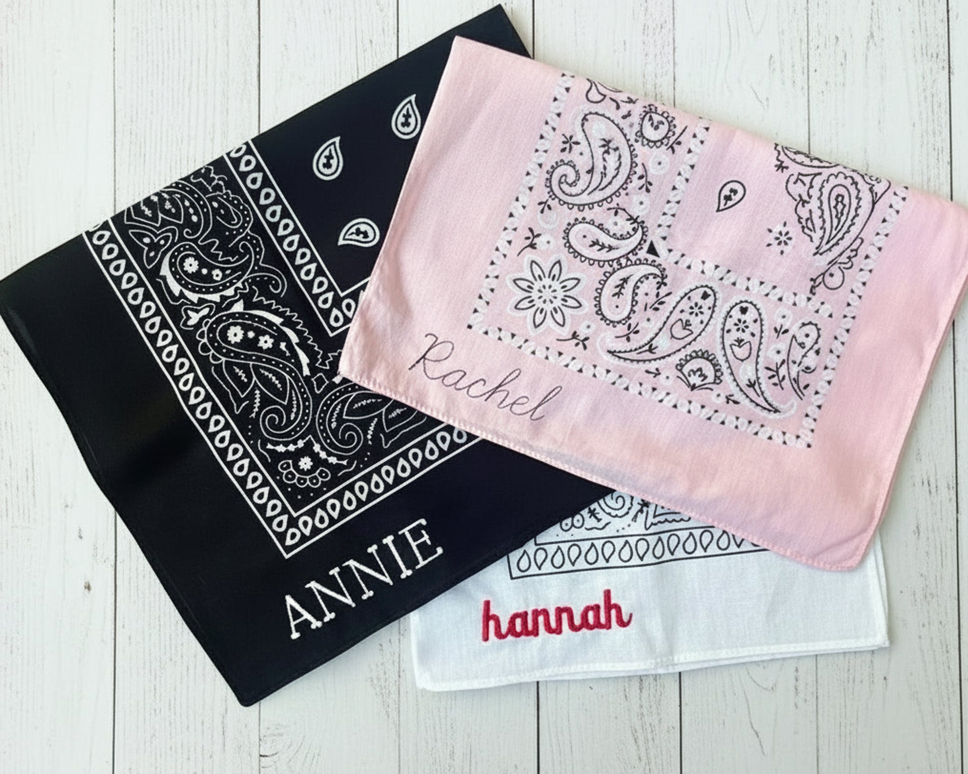 Three folded cotton bandanas in black, pink, and white featuring personalized names "ANNIE," "Rachel," and "hannah" in various embroidery styles.