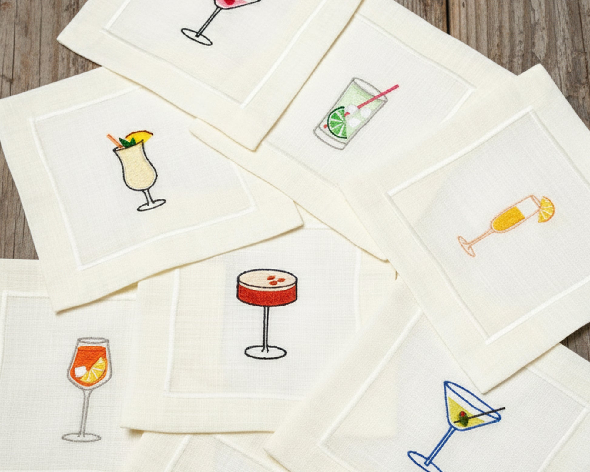 An assorted collection of 100% cotton beverage napkins featuring vibrant embroidered icons of various cocktails like an Aperol Spritz, Espresso Martini, and Champagne.